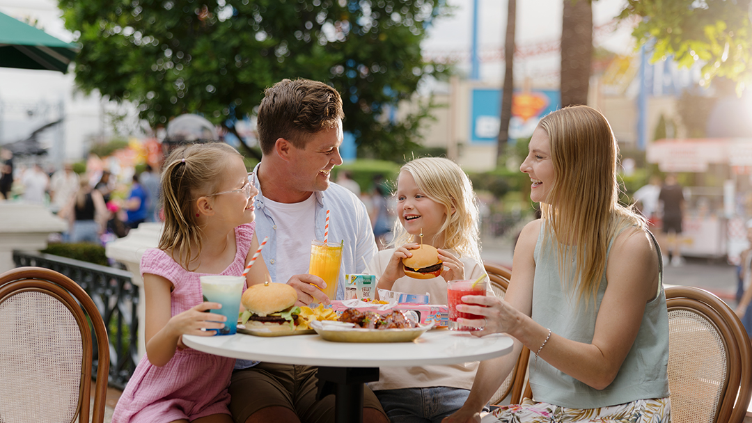 A family of four sits at an outdoor café table, smiling and enjoying burgers and colorful drinks. The parents and two young daughters appear happy, surrounded by a lively outdoor setting.