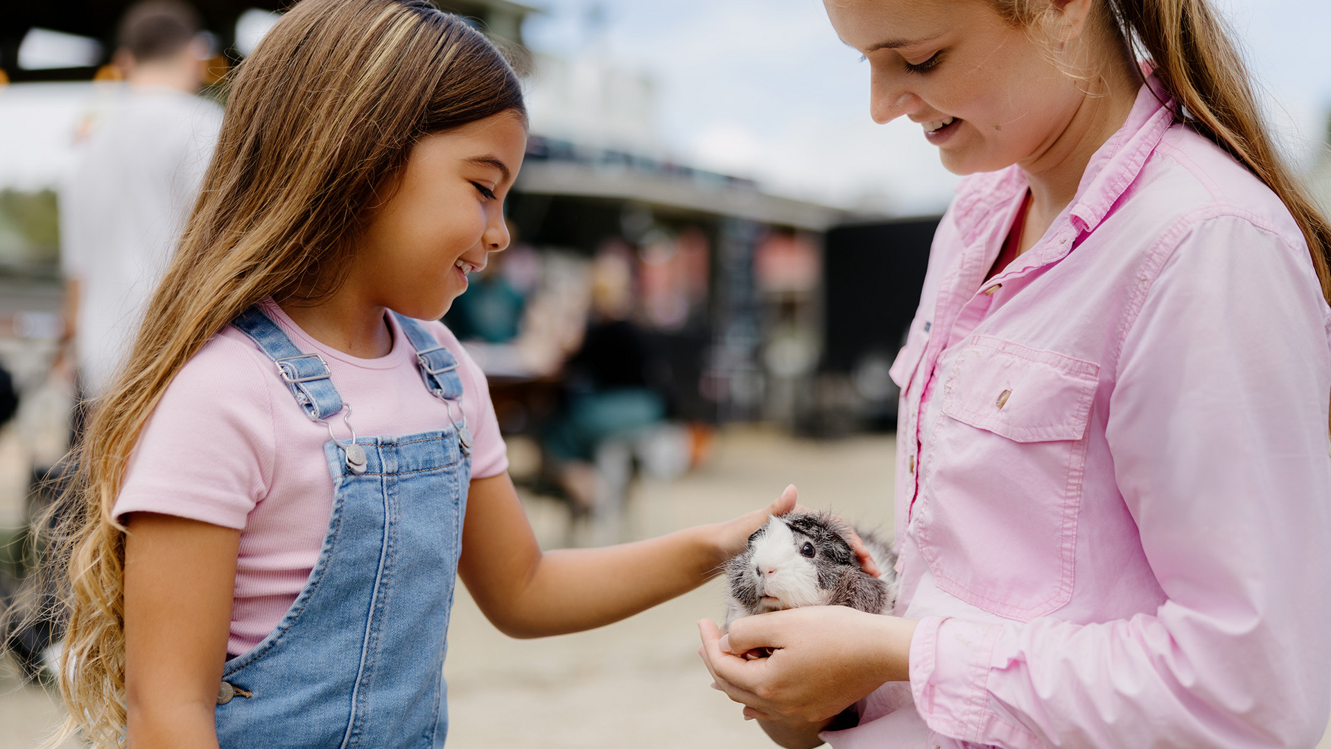 Two girls smile as one gently pets a small animal held by the other. They are outdoors, dressed casually, and appear to be enjoying their interaction with the animal.