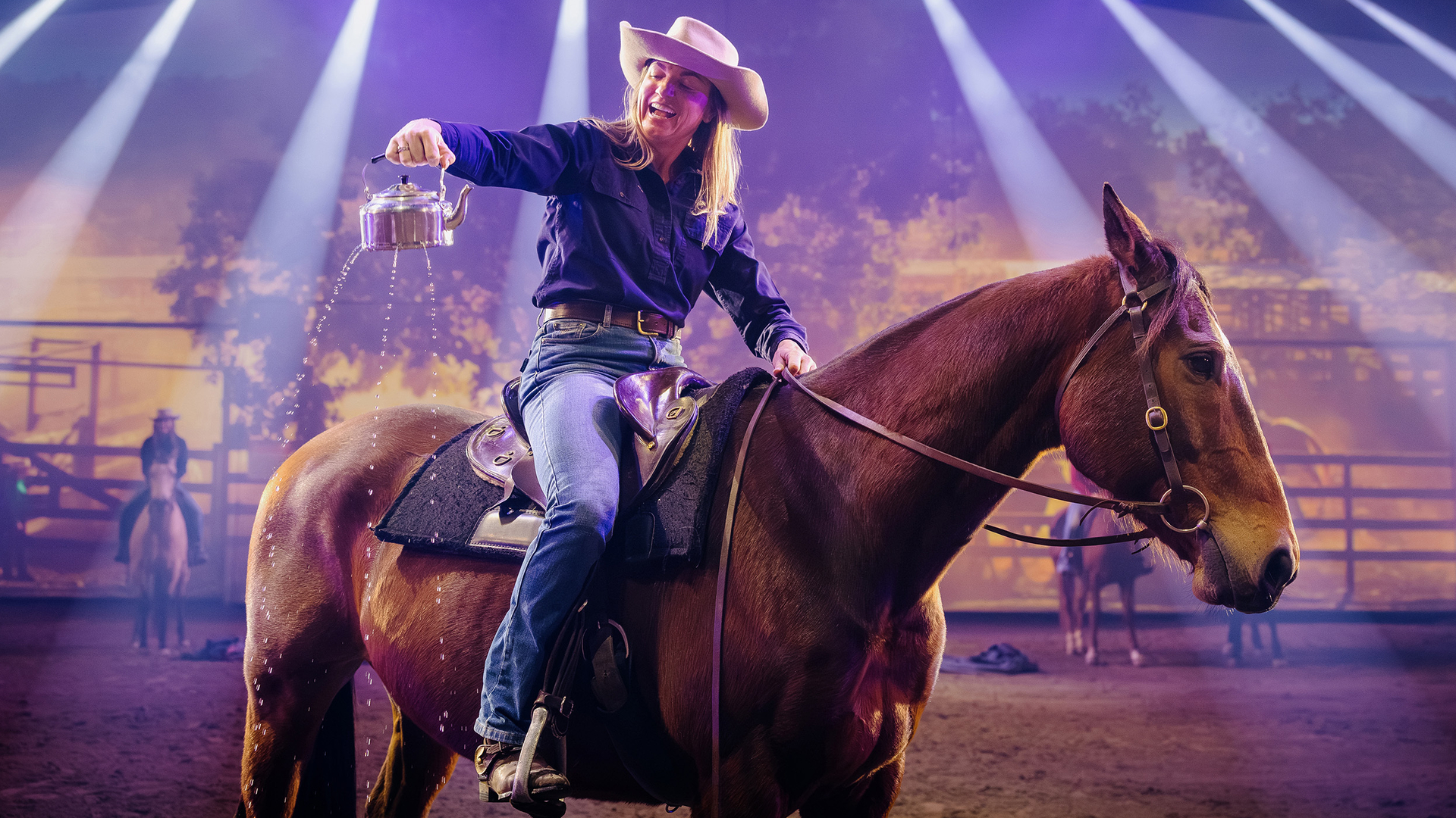 A woman in a cowboy hat sits on a brown horse, pouring water from a metal kettle. She is under bright stage lights with a rural backdrop and other riders blurred in the background.