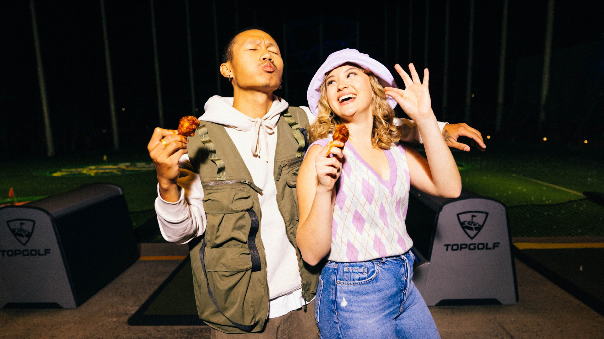 Two people stand together at a Topgolf venue, smiling and holding chicken wings. The man on the left wears a hoodie and vest, while the woman on the right wears a purple hat and patterned top. Golf bays and targets are visible behind them.