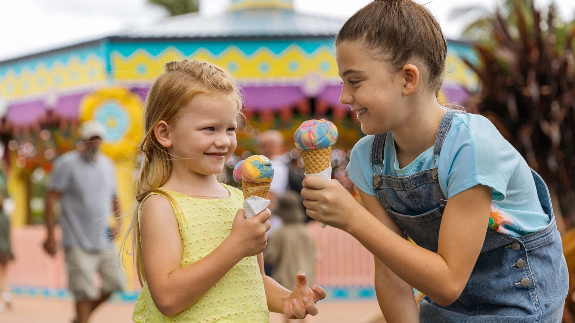 Two young girls smiling and holding colorful ice cream cones outdoors, with a festive building and people blurred in the background.