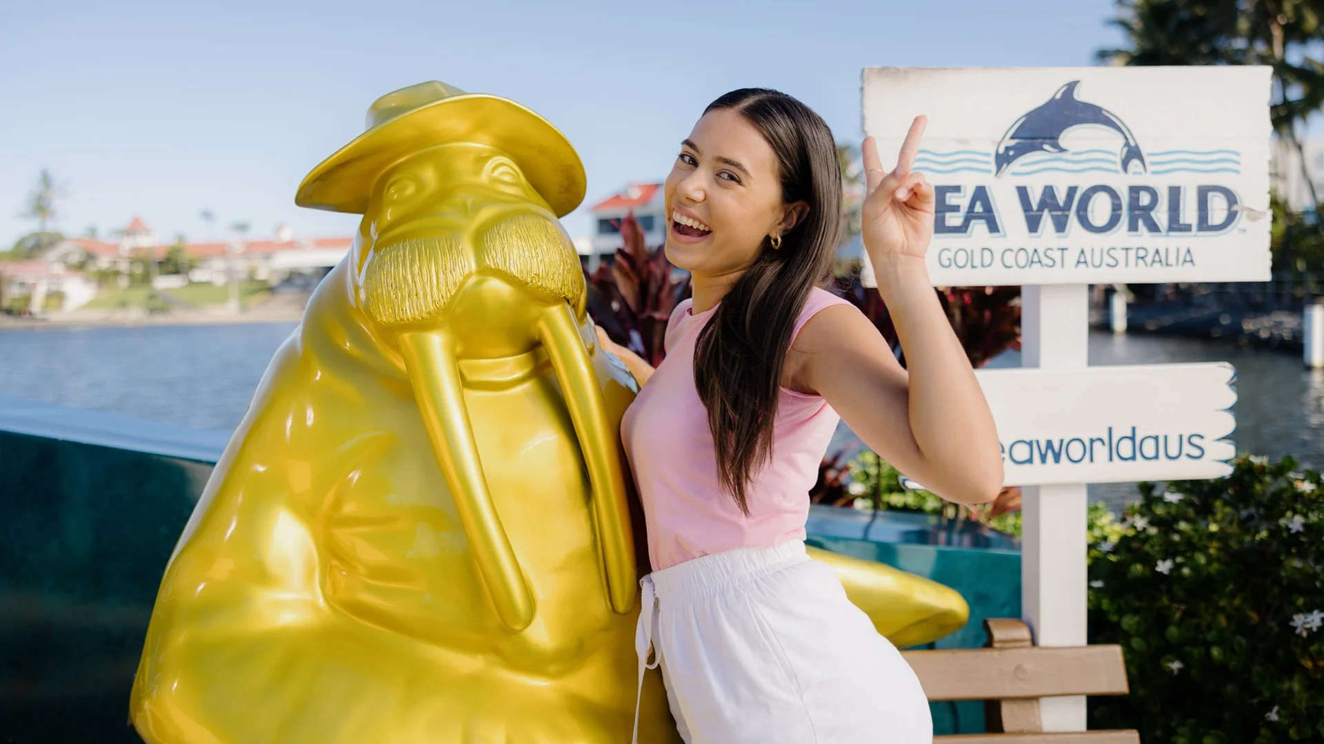 A woman poses, smiling and making a peace sign next to a gold walrus statue at Sea World Gold Coast Australia.
