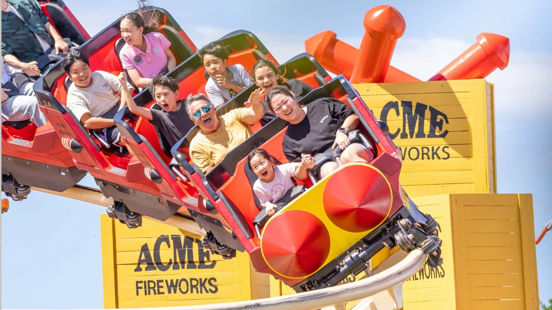 People are riding a roller coaster with yellow ACME Fireworks signage in the background. Some are smiling and raising their hands in excitement.