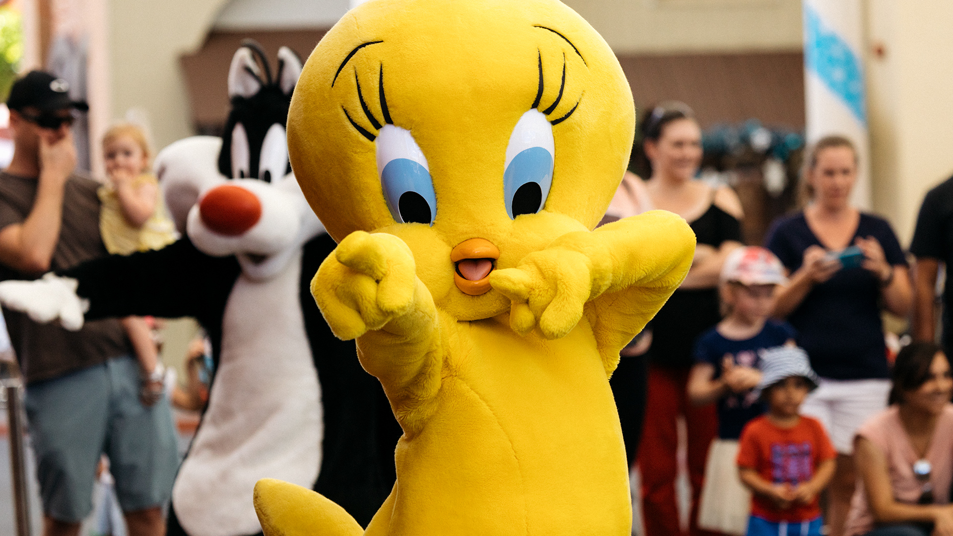 A person in a large Tweety Bird costume poses playfully in front of a crowd, with someone dressed as Sylvester the Cat standing behind. Onlookers, including children and adults, watch and take photos in the background.