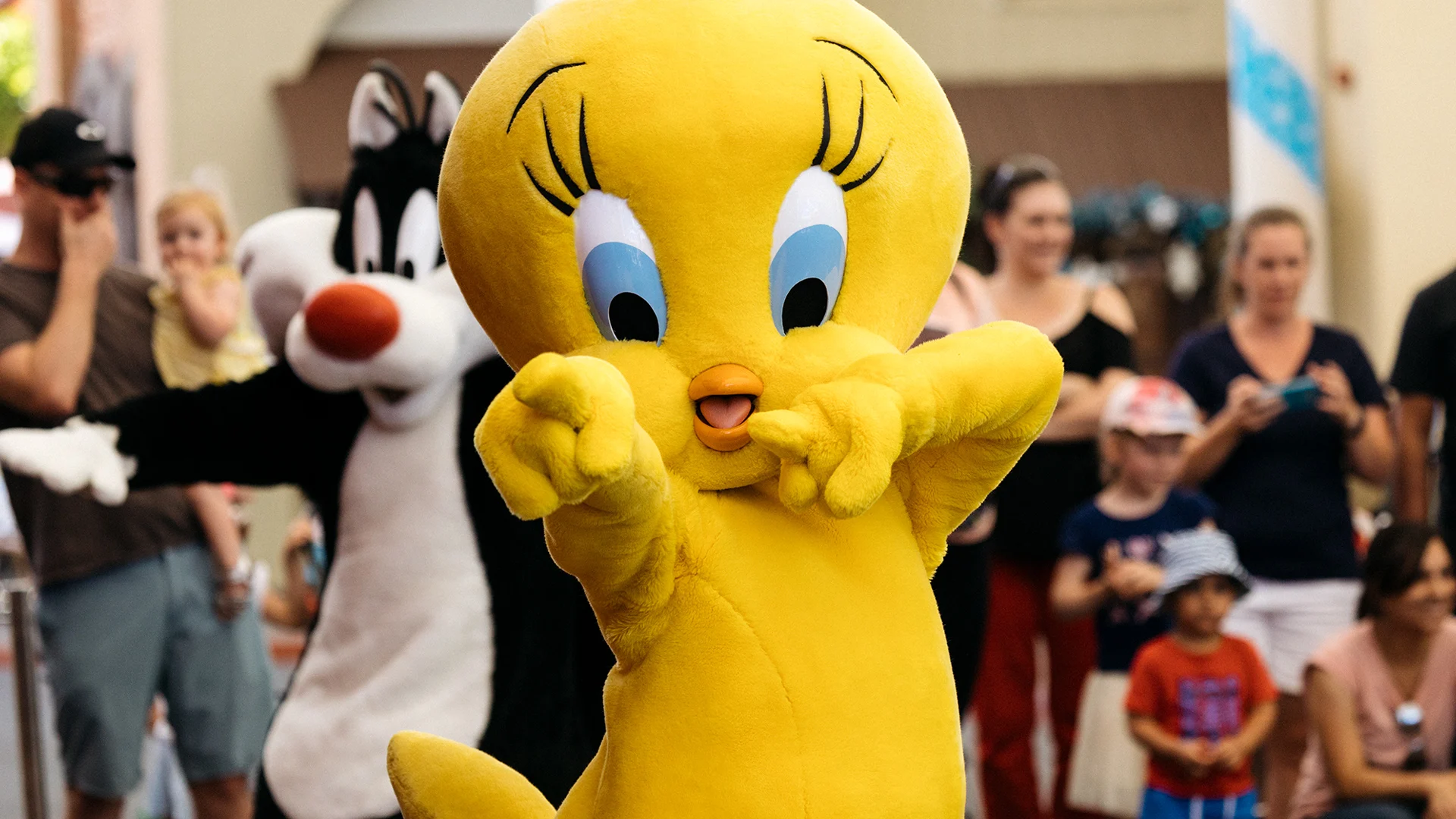 A person in a large Tweety Bird costume poses playfully in front of a crowd, with someone dressed as Sylvester the Cat standing behind. Onlookers, including children and adults, watch and take photos in the background.