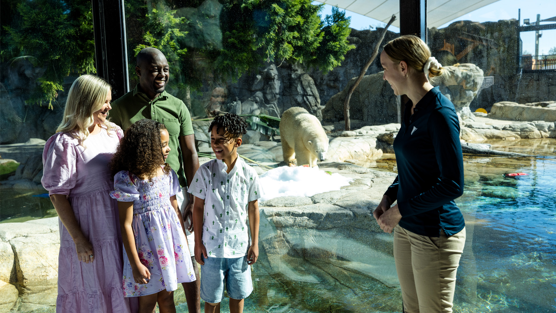 A family of four stands with a zoo staff member near a glass enclosure, watching a polar bear by the water in a zoo exhibit.