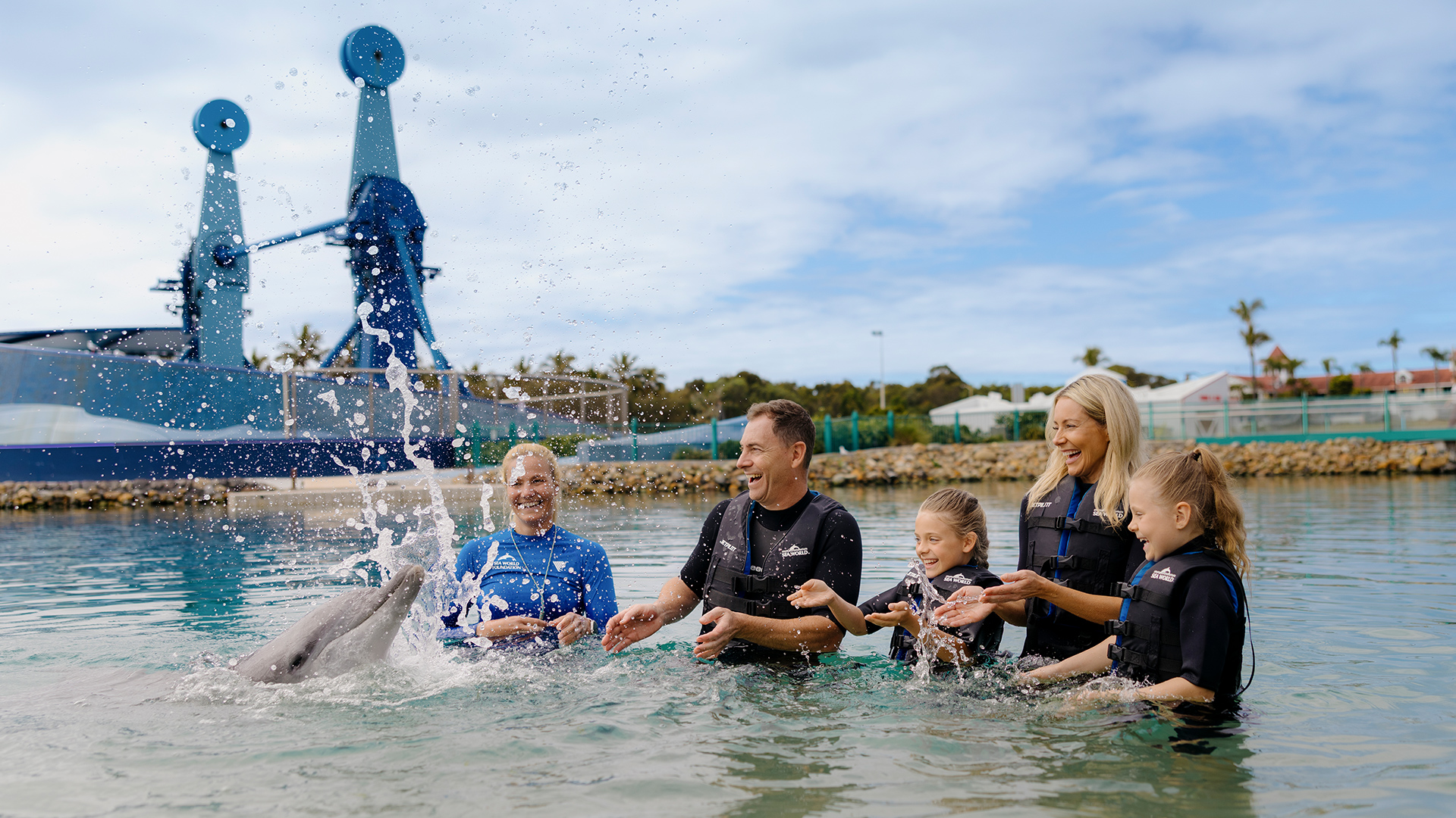A smiling family stands in shallow water at a marine park, interacting with a dolphin that splashes water at them. Palm trees, structures, and a blue sky are visible in the background.