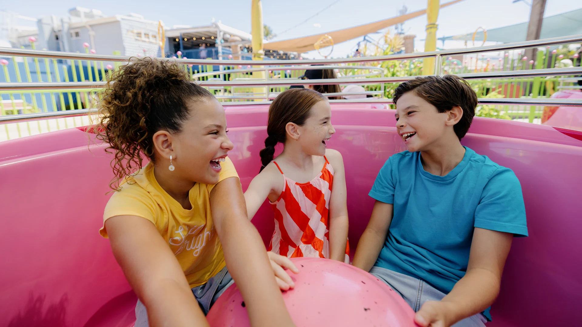 Three children sit in a pink amusement park ride, smiling and laughing together on a sunny day.