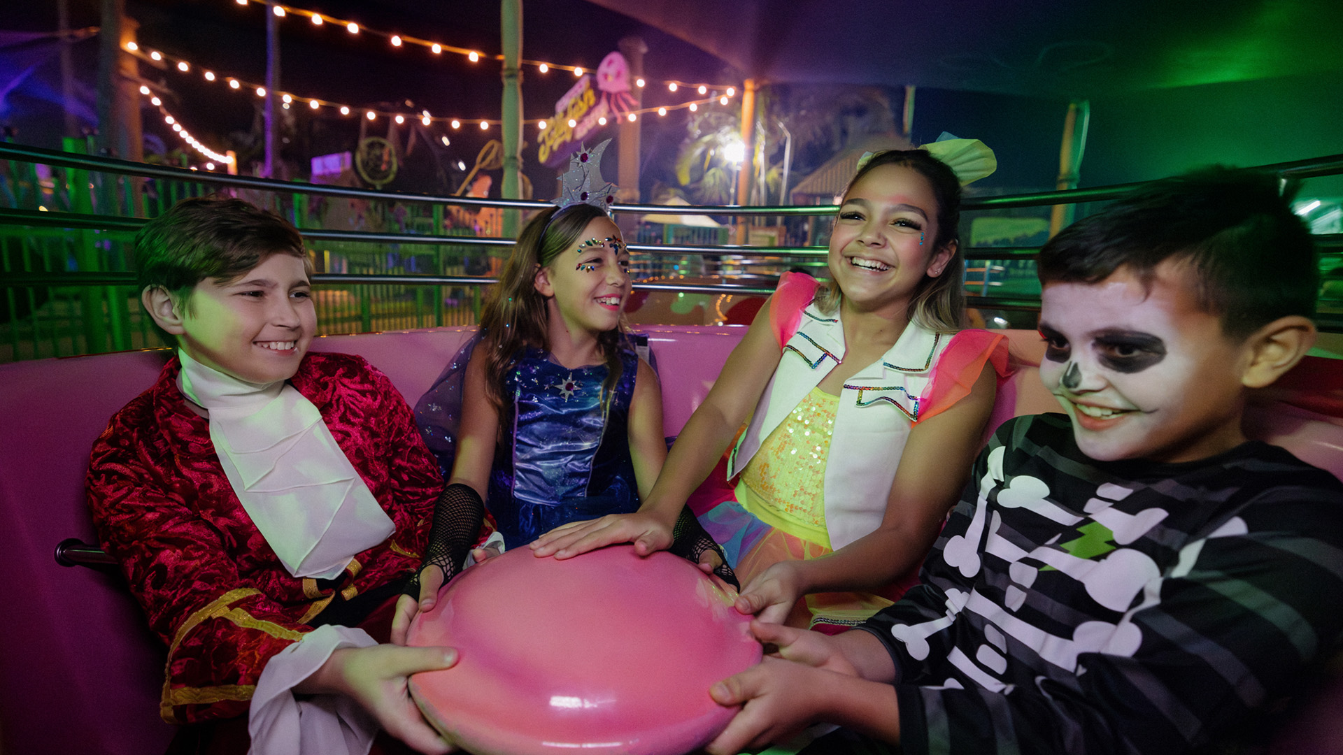 Four children in colorful costumes, including a skeleton and royalty, smile and laugh while spinning on an amusement park ride under festive lights at night.