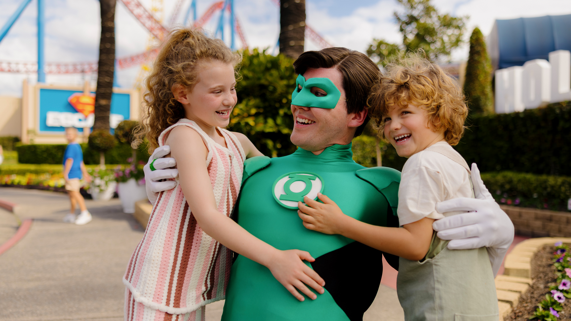 A person dressed as the superhero Green Lantern hugs two smiling young children at an amusement park, with roller coasters and the Superman logo visible in the background.