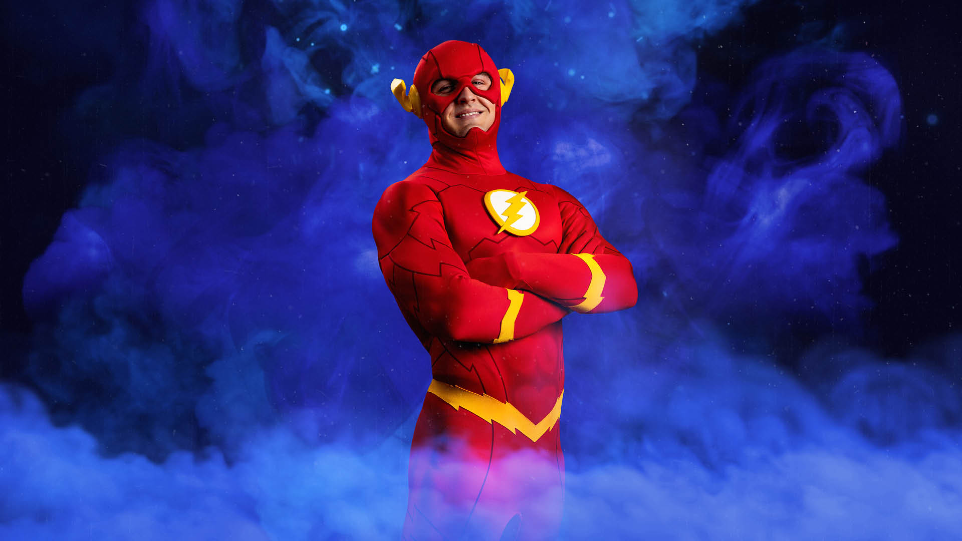 Person dressed as the Flash superhero stands with arms crossed, smiling, in front of a purple smoky background.