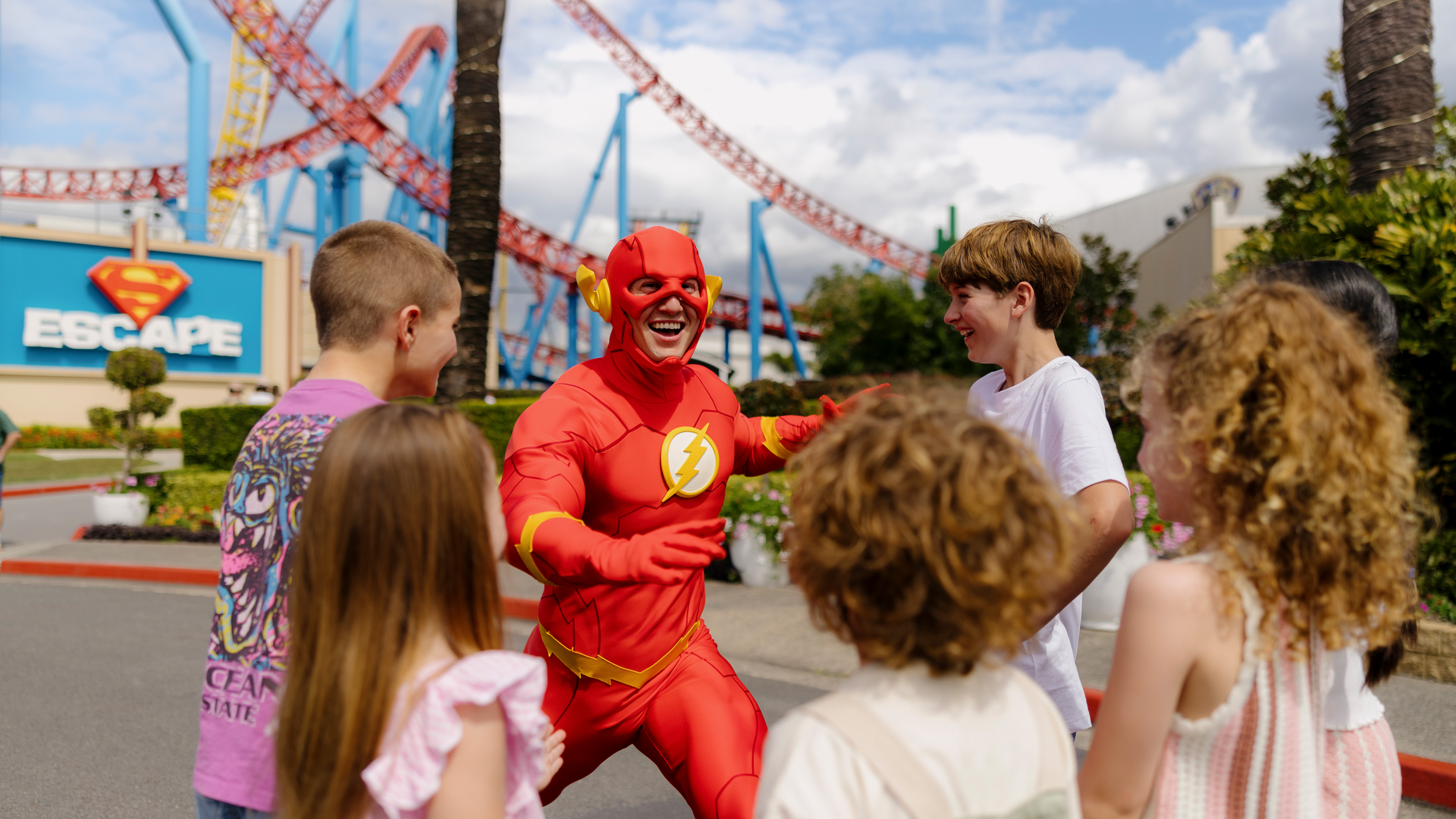 A person dressed as the Flash greets a group of smiling children at an amusement park, with a Superman-themed ride and red roller coaster tracks visible in the background.