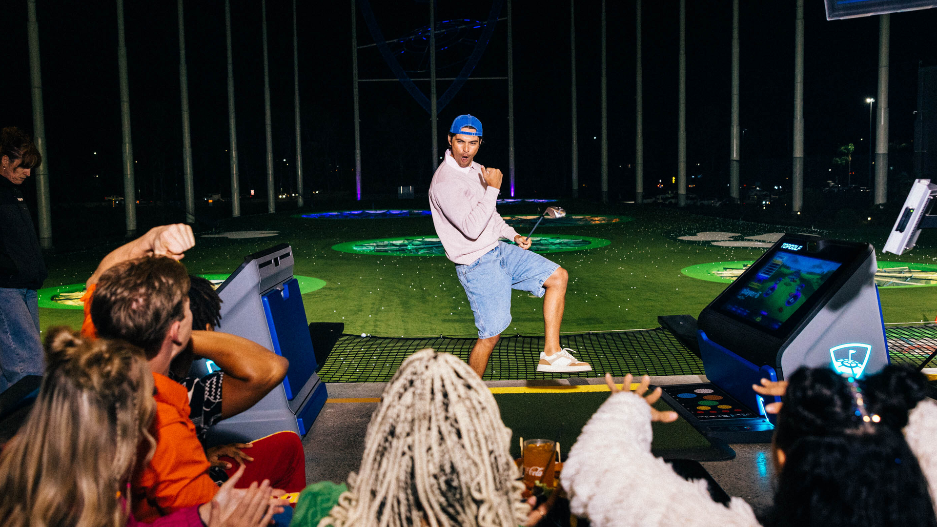 A man in a pink shirt and blue hat strikes a playful pose with a golf club at a nighttime golf range, while a group of people cheer him on from behind. Bright lights and colorful targets are visible in the background.