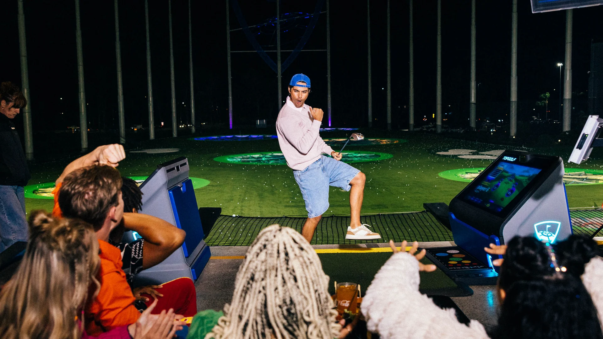 A man in a pink shirt and blue hat strikes a playful pose with a golf club at a nighttime golf range, while a group of people cheer him on from behind. Bright lights and colorful targets are visible in the background.