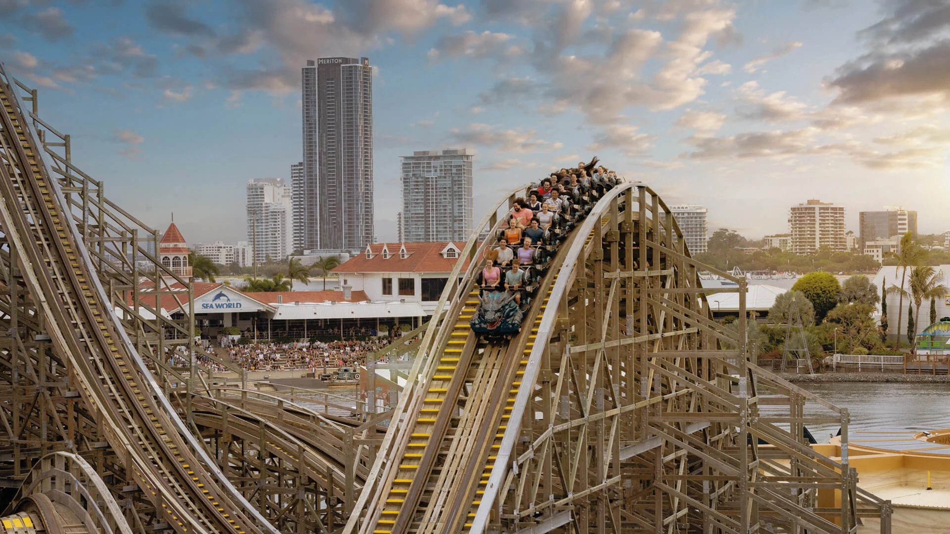 Roller coaster with riders descending a wooden track against a cityscape background of tall buildings and a clear sky.