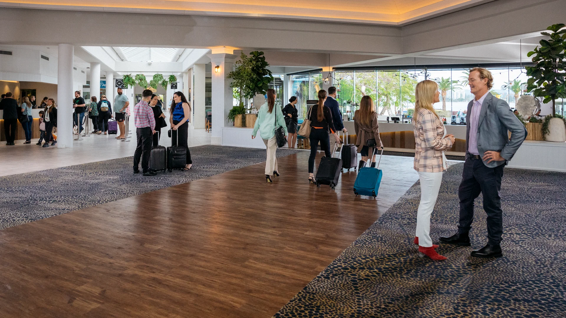 Hotel lobby with people checking in, talking, and pulling suitcases. Large windows let in natural light, and the lobby is decorated with plants and contemporary furnishings.