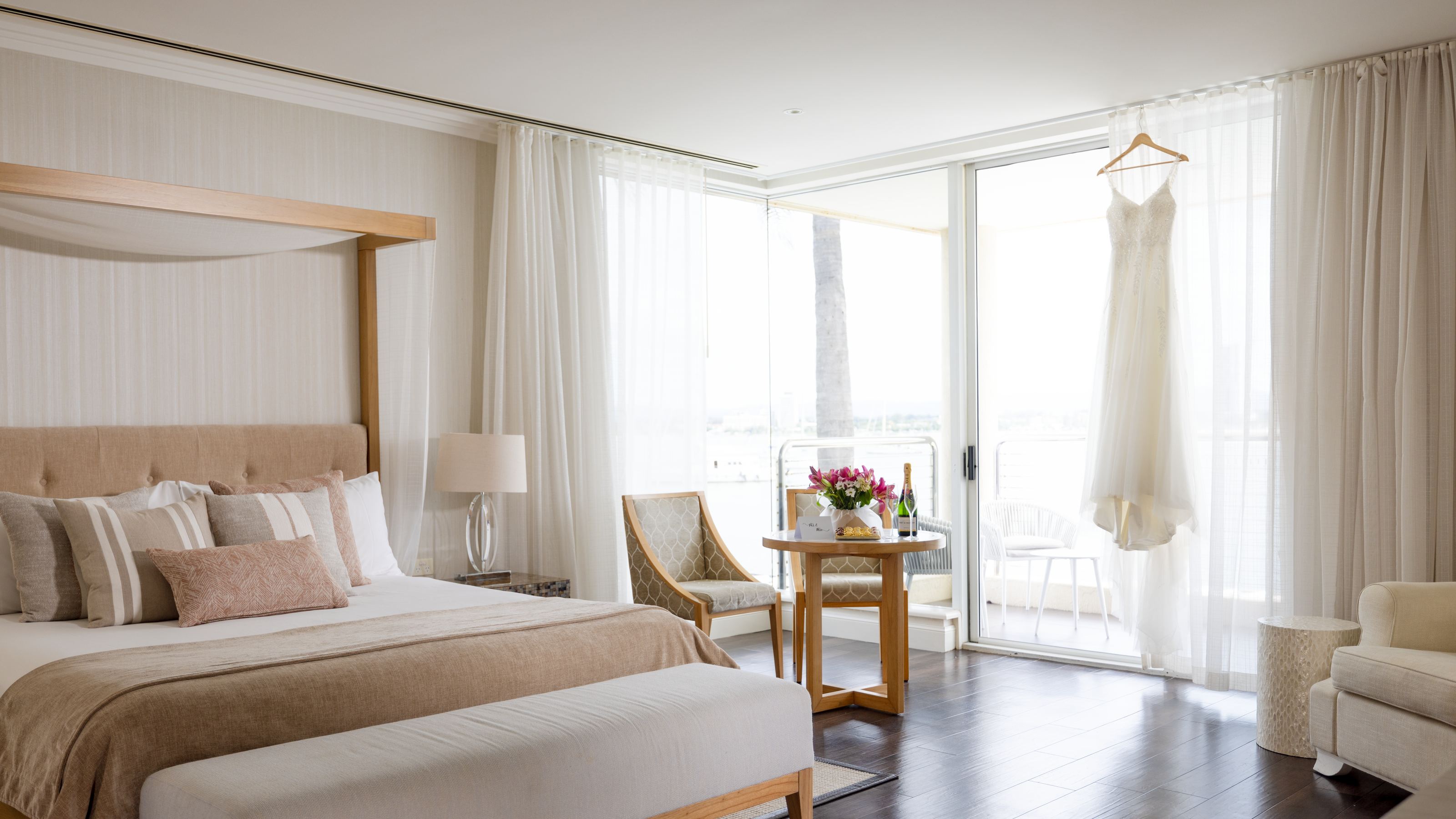 Bright hotel room with a large bed, wooden canopy, small round table with flowers, two chairs, and a white dress hanging by a window. The room features sheer curtains and a light color scheme.