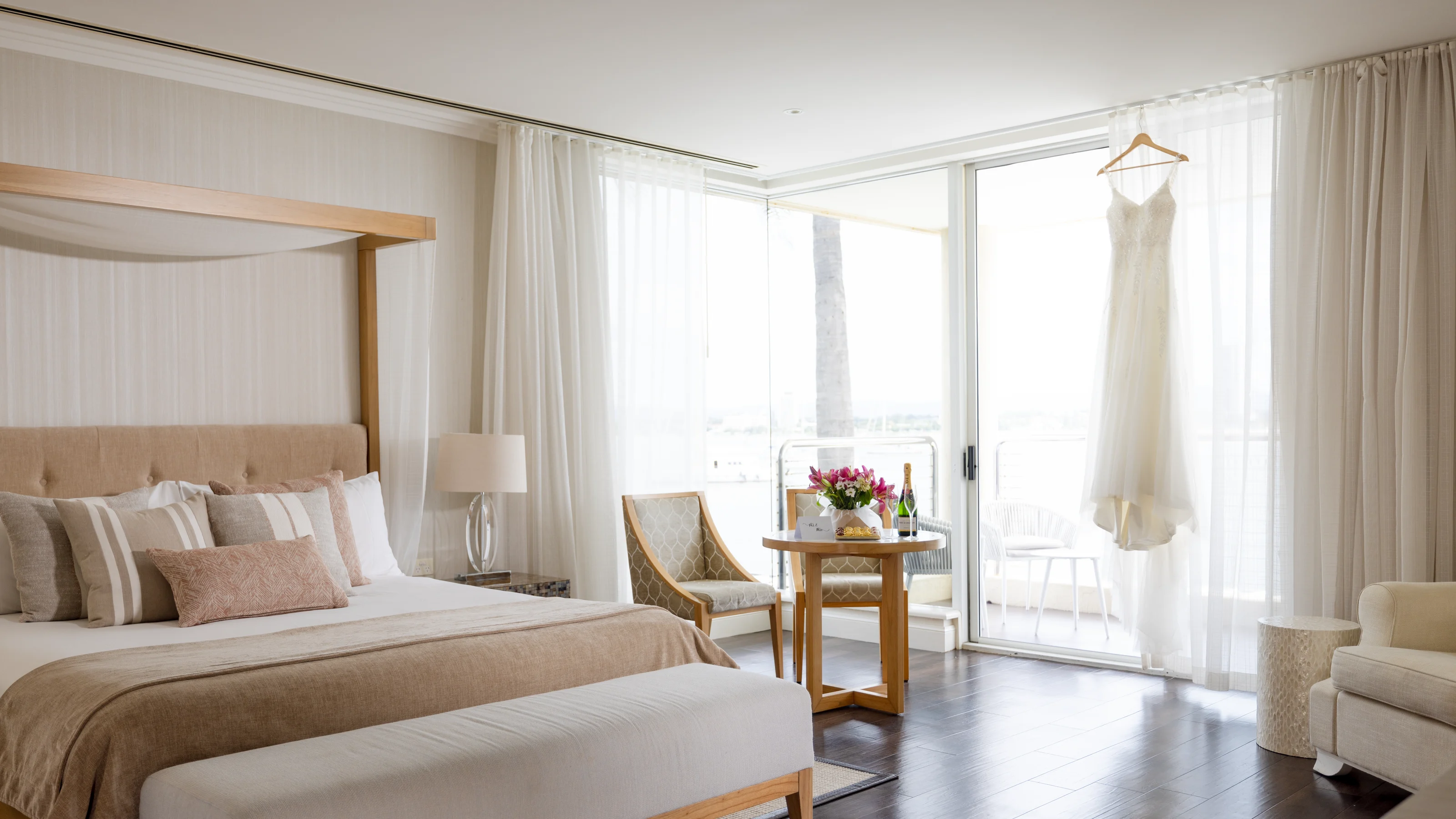 Bright hotel room with a large bed, wooden canopy, small round table with flowers, two chairs, and a white dress hanging by a window. The room features sheer curtains and a light color scheme.