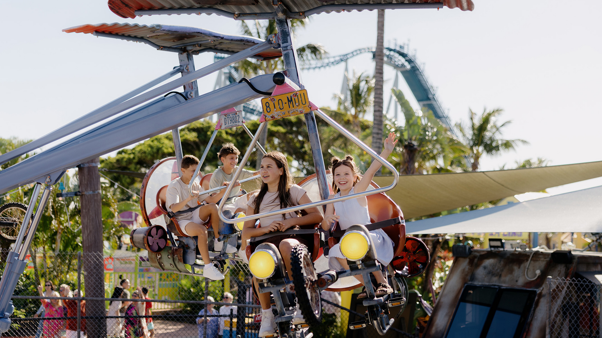 Four children are riding a flying amusement park ride shaped like airplanes. Two of them are smiling and waving, surrounded by palm trees and colorful attractions on a sunny day.