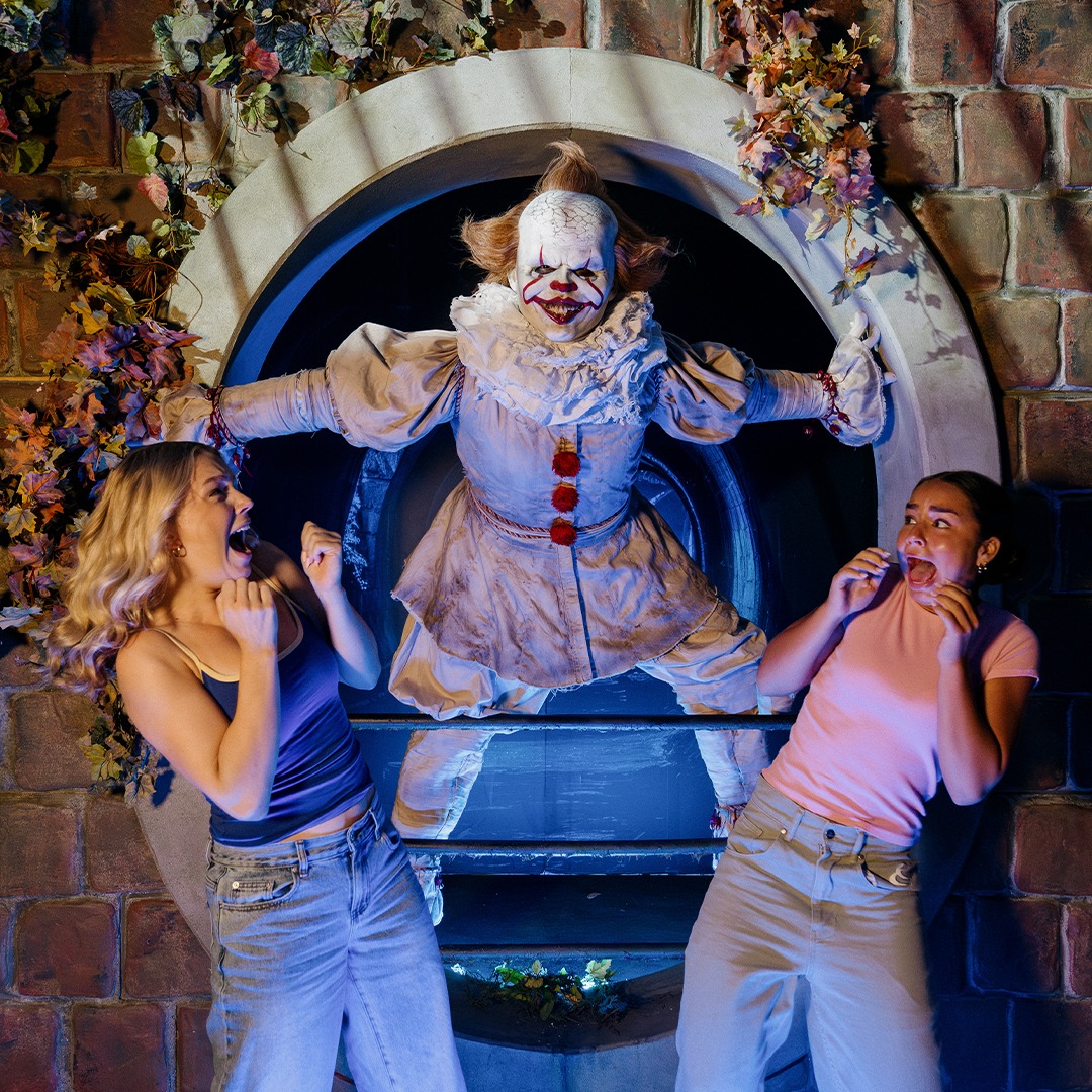 Two frightened women react to a clown in a sewer opening, dressed in a white costume with red accents, against a brick wall with foliage.