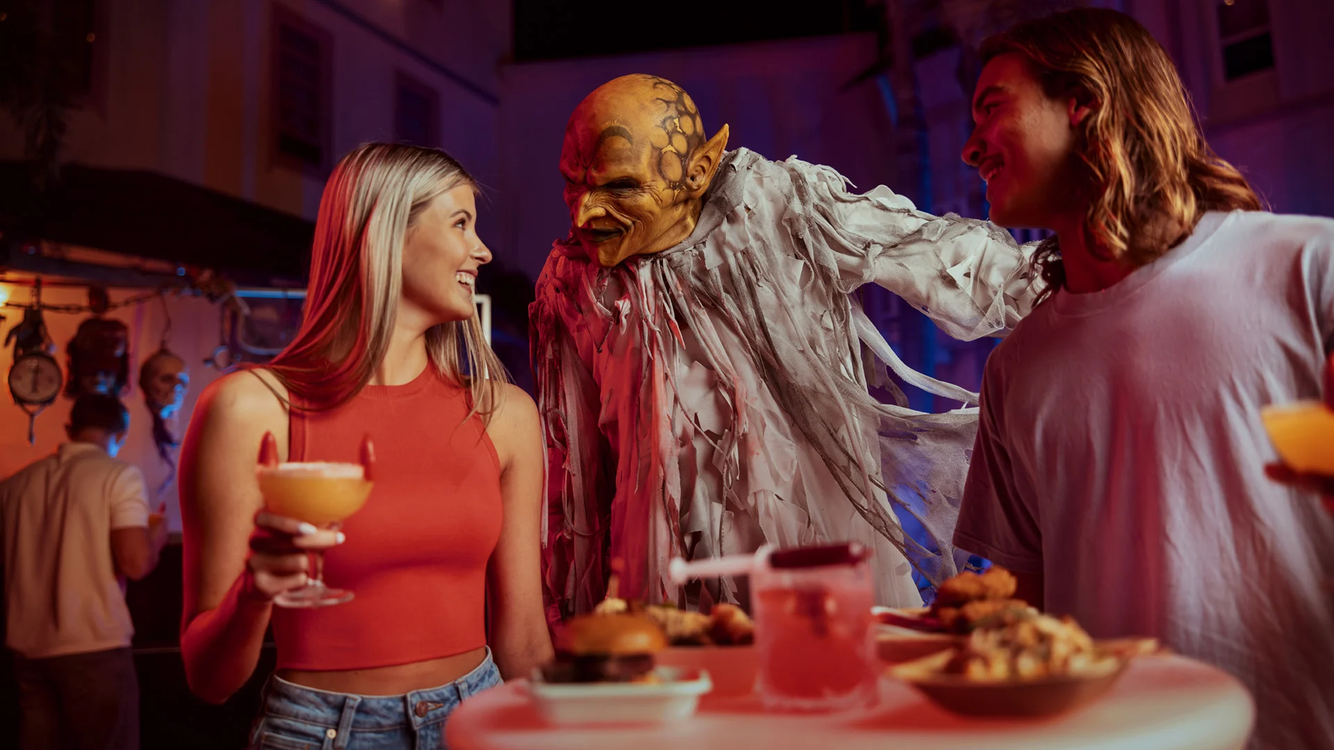 Two people smile and talk at a party table with food and drinks while a person in a spooky goblin costume stands behind them, creating a fun and festive atmosphere.