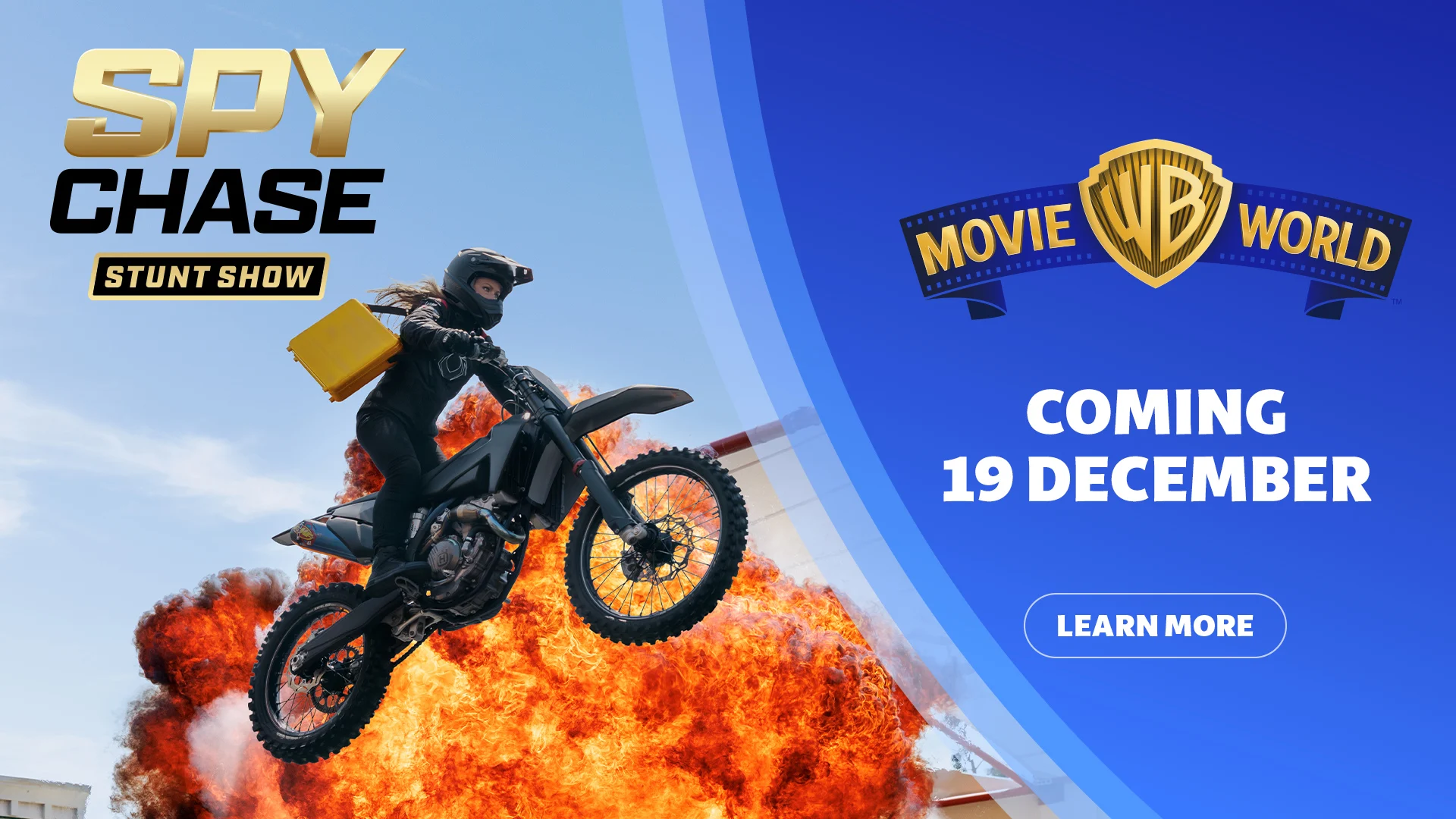 A motorcycle stunt rider in black jumpsuit and helmet leaps in front of a fiery explosion. Text reads, "Coming 19 December. Spy Chase Stunt Show. Movie World." Warner Bros and Village Roadshow Theme Parks logos are visible.