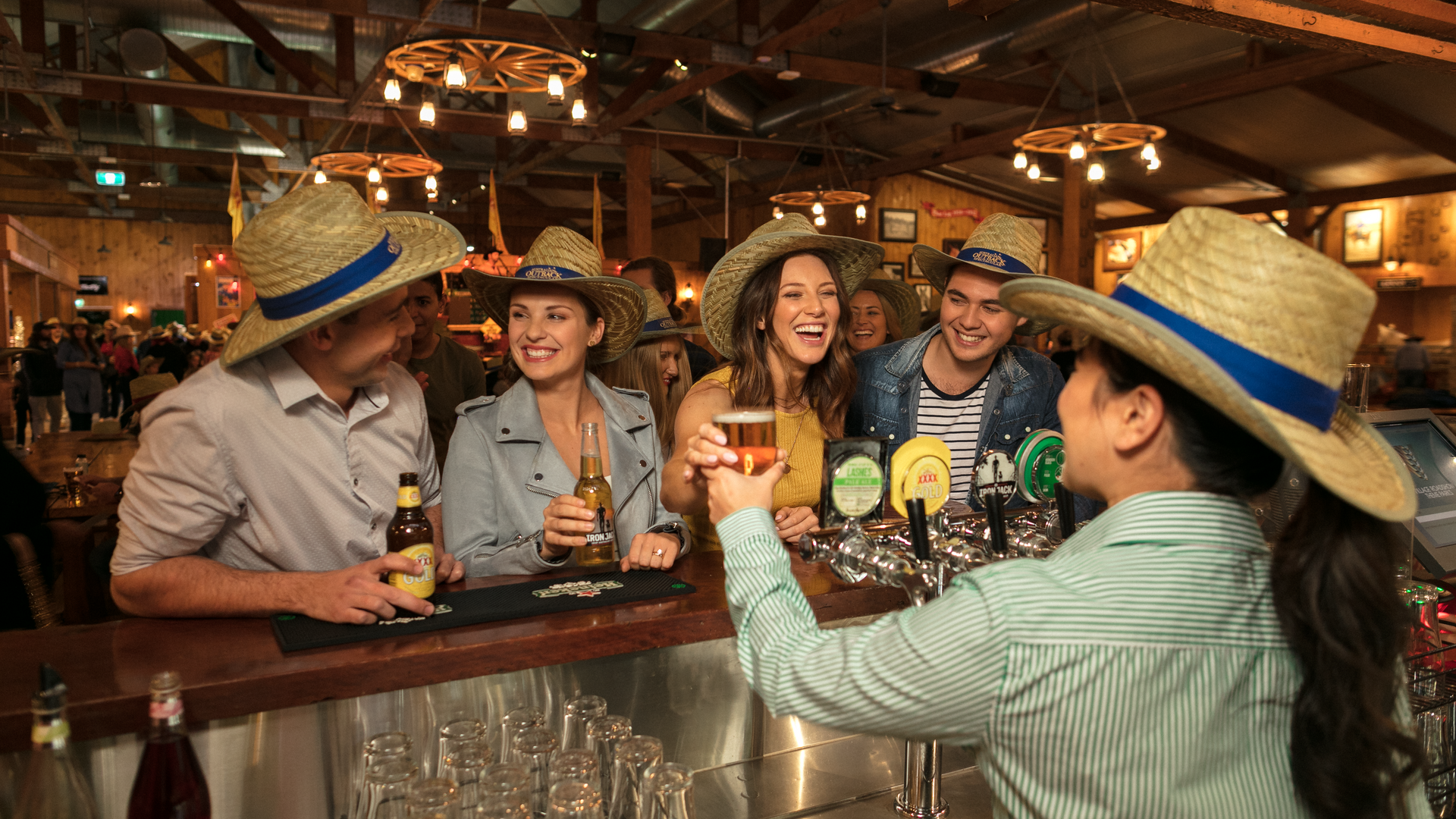 A group of people wearing straw hats laugh and chat at a bar counter, drinks in hand, while a bartender in a similar hat serves them in a warmly lit, rustic bar.