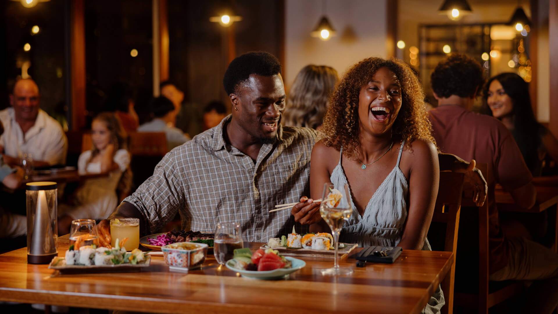 A couple is enjoying sushi and drinks at a Japanese restaurant, smiling and laughing. The table is set with plates of food and glasses. Other diners are visible in the background.