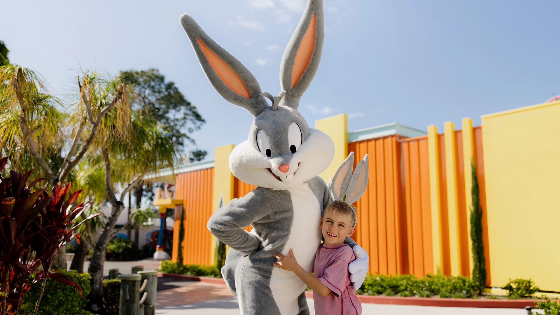 A young boy hugs Bugs Bunny outside, near colorful orange and yellow buildings with palm trees and greenery under a clear blue sky.