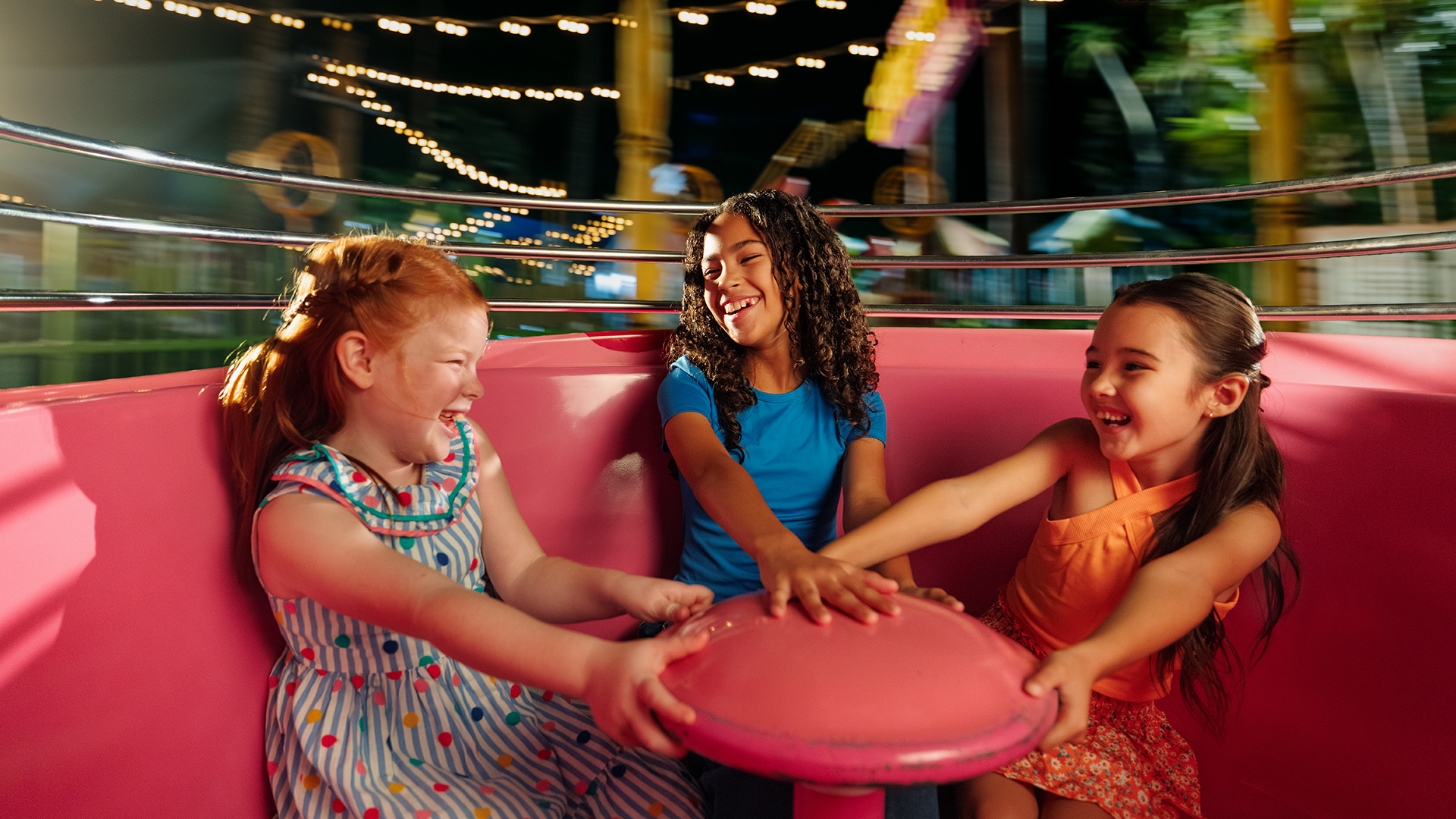 Three young girls laugh and hold onto the center of a spinning pink teacup ride at an amusement park, surrounded by colorful lights and a blurred background.