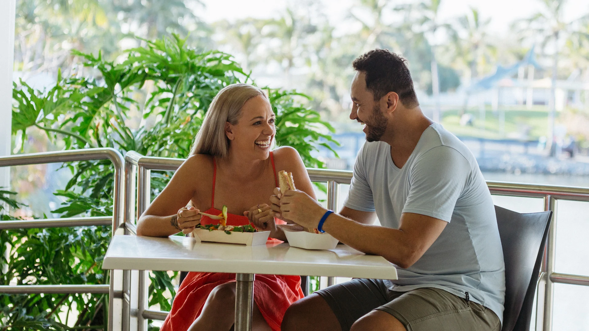 A woman and a man sit across from each other at an outdoor table, smiling and eating sandwiches, with greenery and trees in the background.