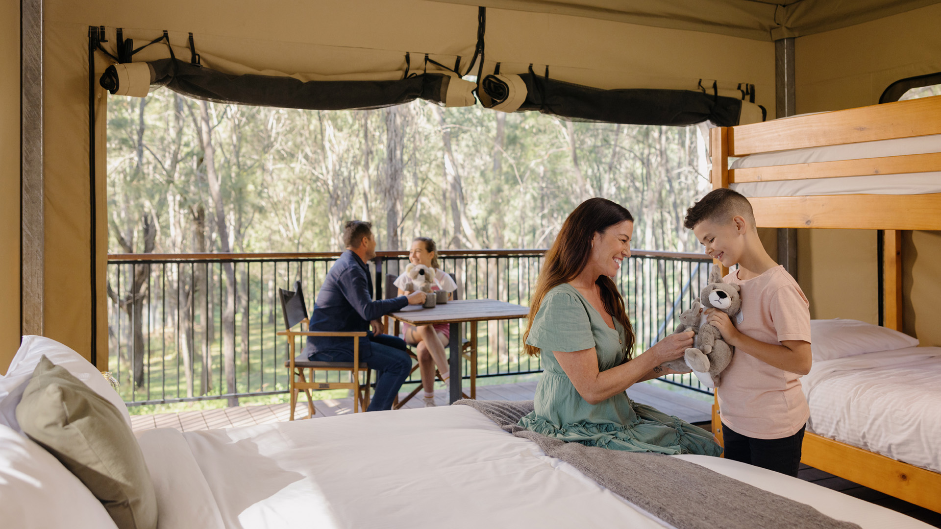 A woman and child sit by a bunk bed inside a tent, holding a stuffed animal, while two people sit at a table on the deck outside, surrounded by trees.