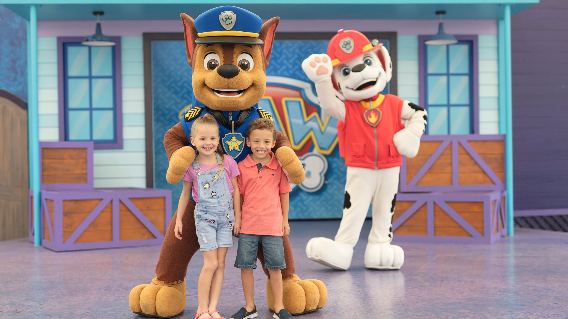 Two children pose with costumed characters, a dog in a police uniform and a dog in a firefighter outfit, on a colorful stage.