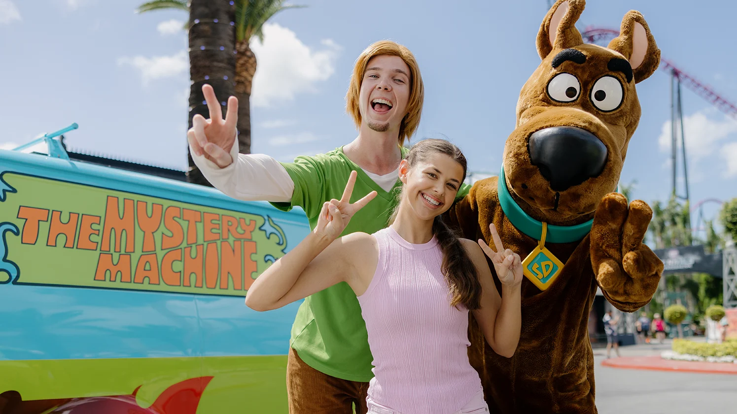 A woman poses smiling with Shaggy and Scooby-Doo characters in front of the colorful Mystery Machine van at an outdoor theme park, all making peace signs with their hands.