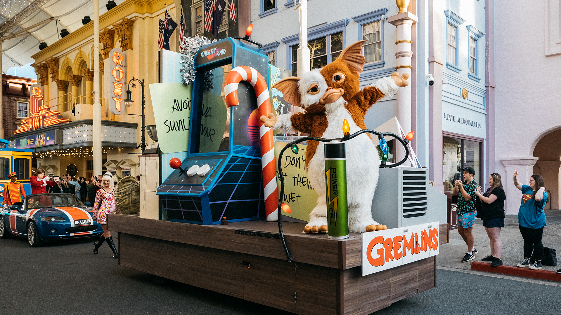 A parade float features a Gremlins-themed display with a large Gizmo figure, candy cane decorations, and arcade props. People watch and take photos as the float passes through a city street lined with colorful buildings.
