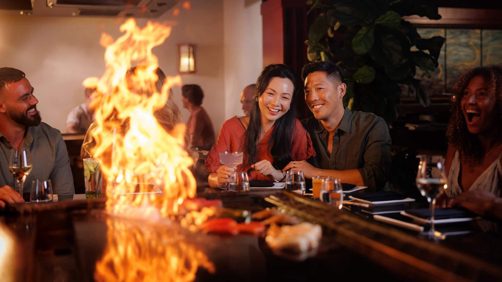 People sitting around a teppanyaki grill with flames rising, enjoying drinks and food in a lively indoor setting.