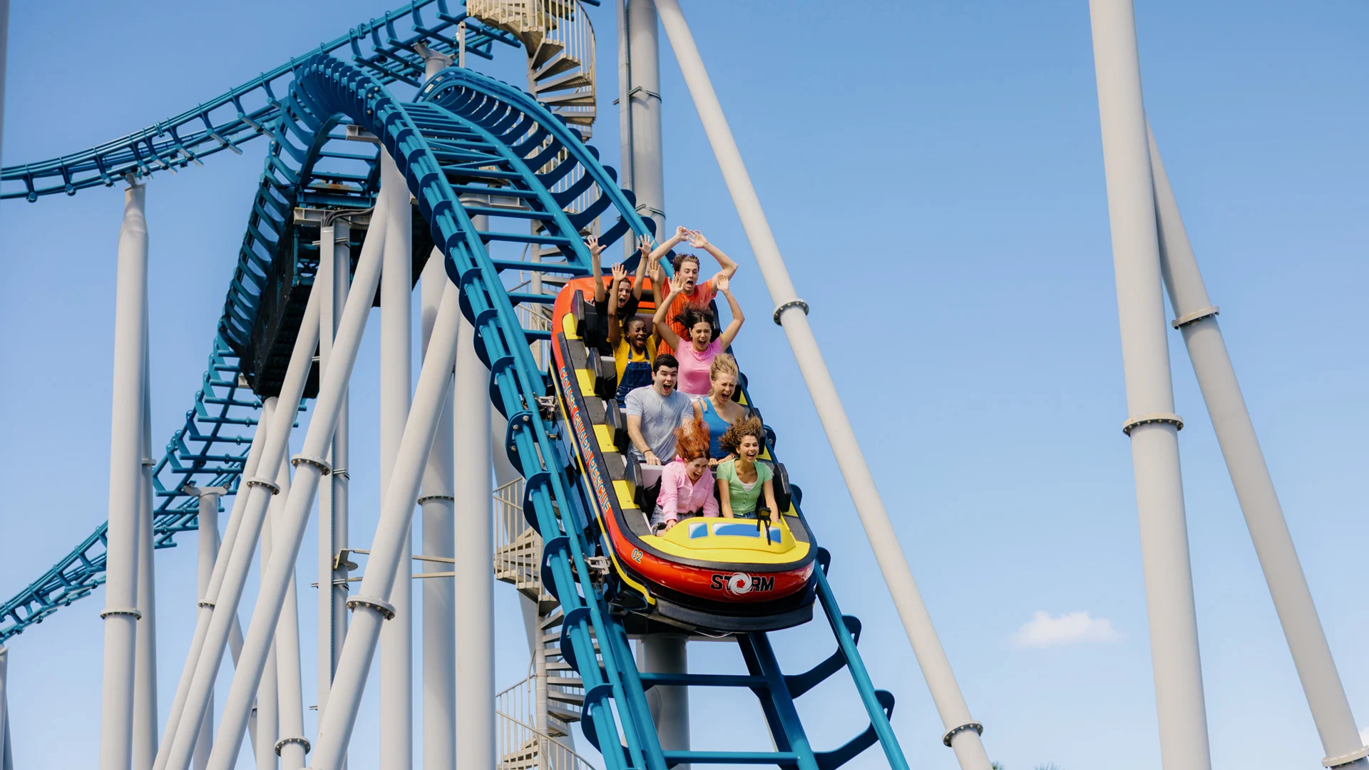 A group of people ride a blue metal roller coaster, some with raised arms, as the car descends from a high point under a clear sky.