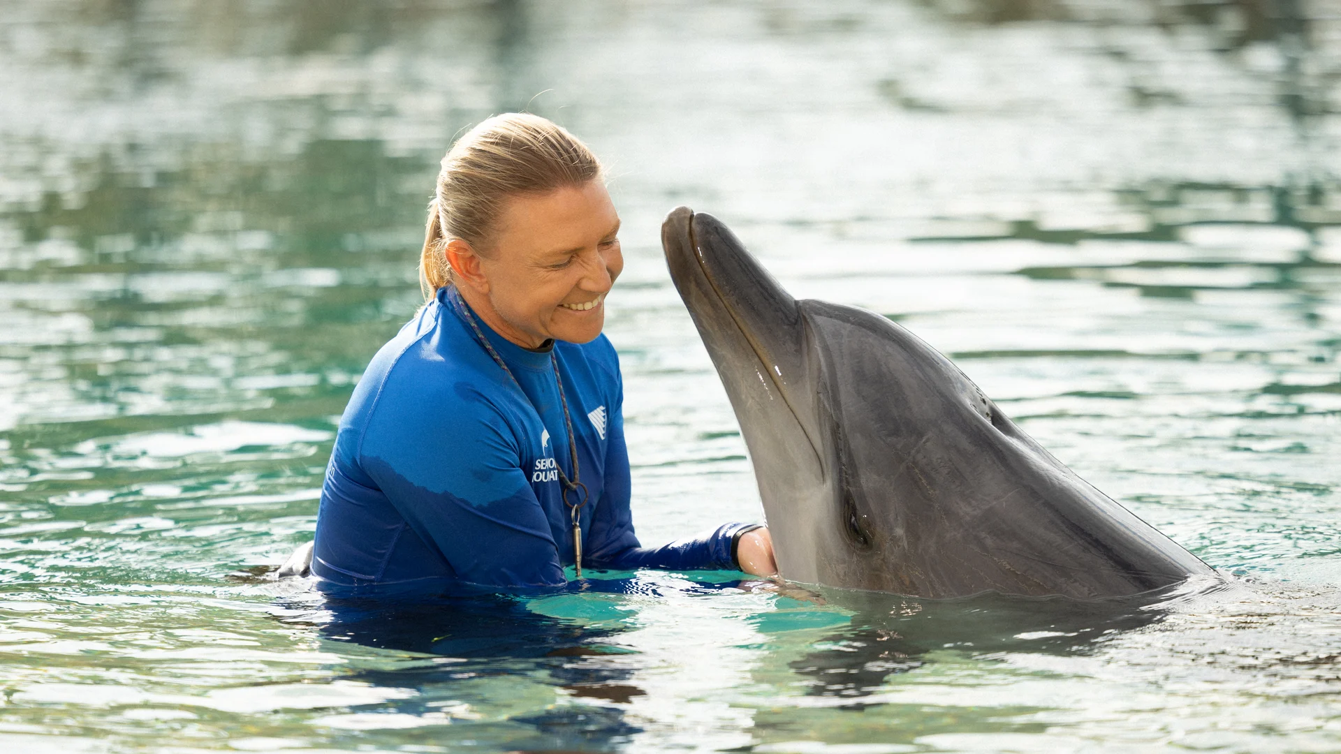 A person in a blue wetsuit stands in the water, smiling and gently holding a dolphin as it lifts its head above the surface.