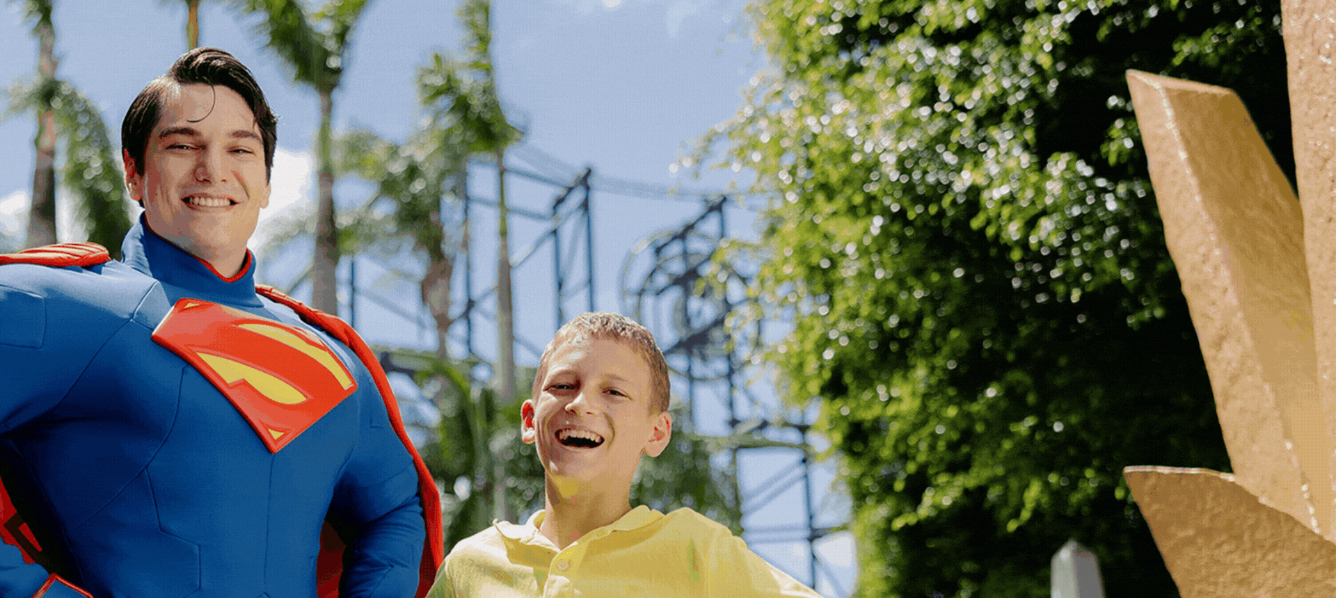A person in a superhero costume stands smiling next to a joyful child outdoors, with trees and a structure in the background.