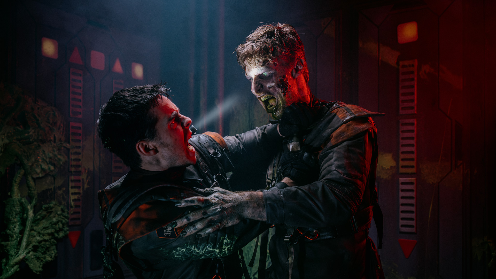 Two people in dark, rugged costumes struggle violently in a dim, industrial setting; one has glowing eyes and zombie-like features, holding the other man by the collar as he tries to fight back.