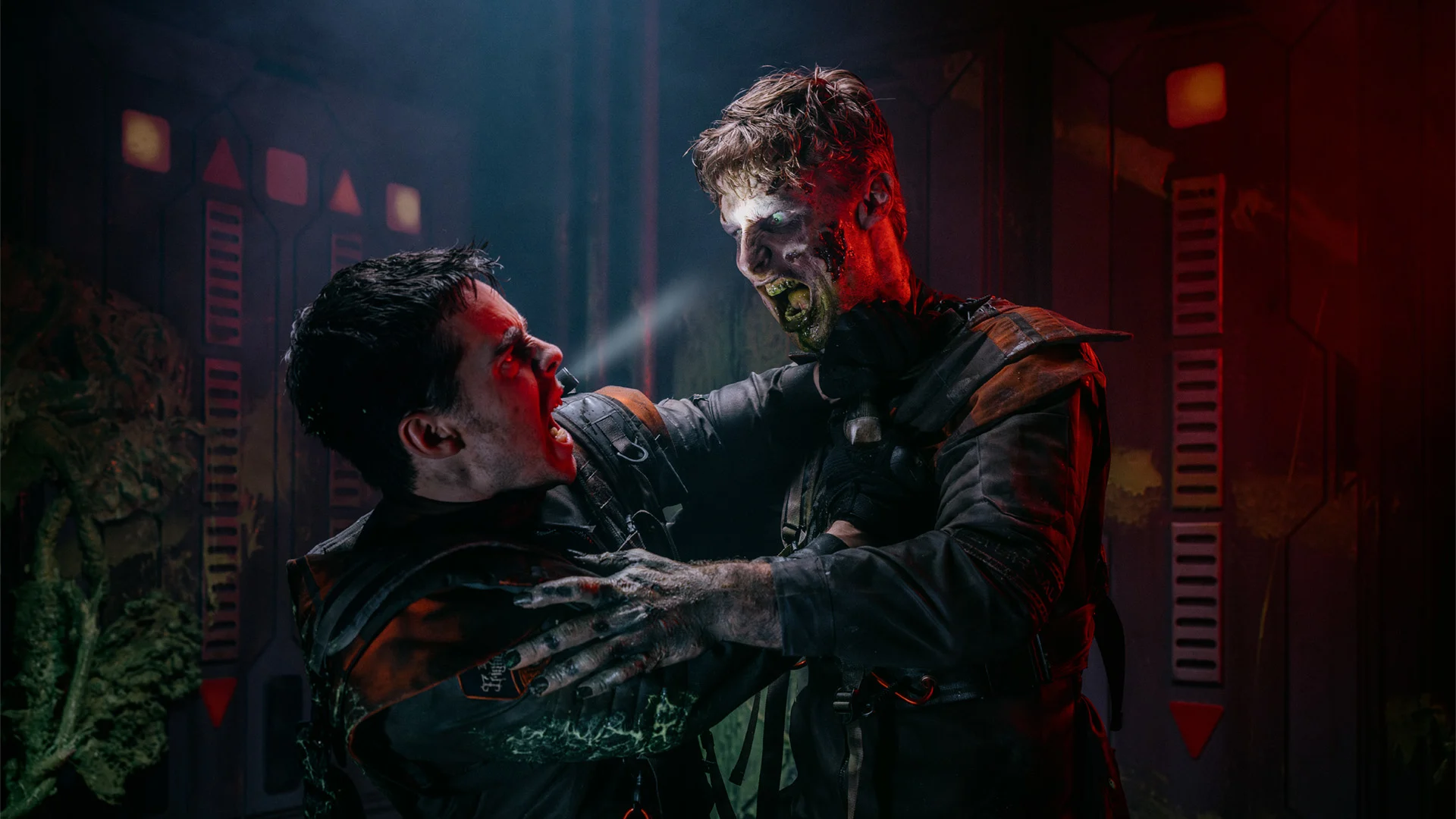Two people in dark, rugged costumes struggle violently in a dim, industrial setting; one has glowing eyes and zombie-like features, holding the other man by the collar as he tries to fight back.