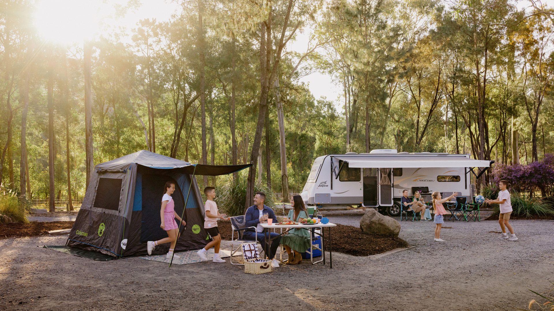 Family camping scene with a tent and RV in a forest. People are sitting and standing around a table with food. Children are playing. Sunlight filters through the trees.