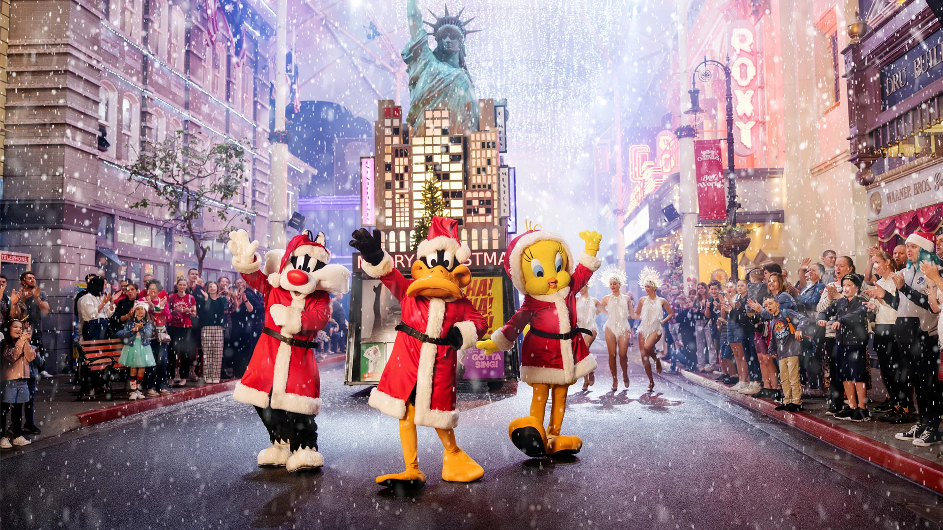Three people in Looney Tunes costumes—Sylvester, Daffy Duck, and Tweety—dressed in Santa outfits, wave and pose during a festive parade with a snowy New York City backdrop and crowds of people applauding.