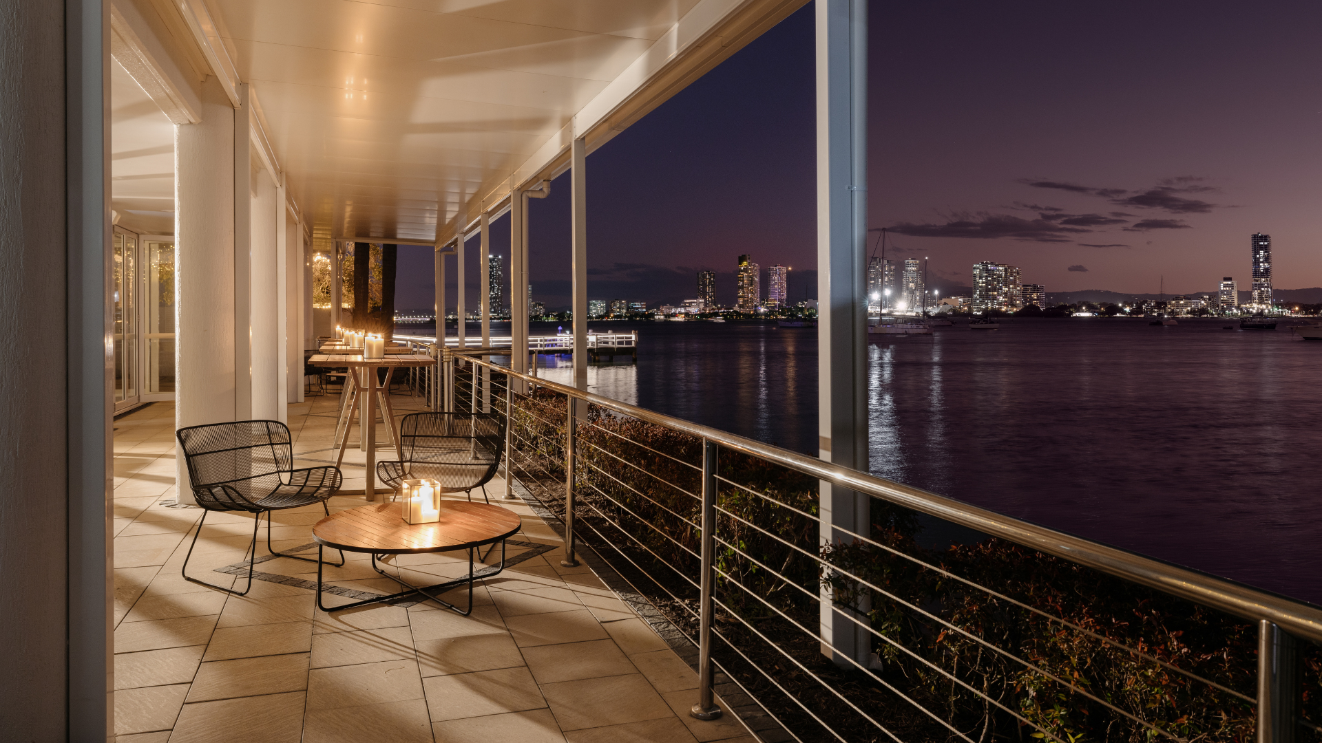 A candlelit patio with chairs and tables overlooks a calm river at night, with city buildings and lights reflecting on the water in the background.