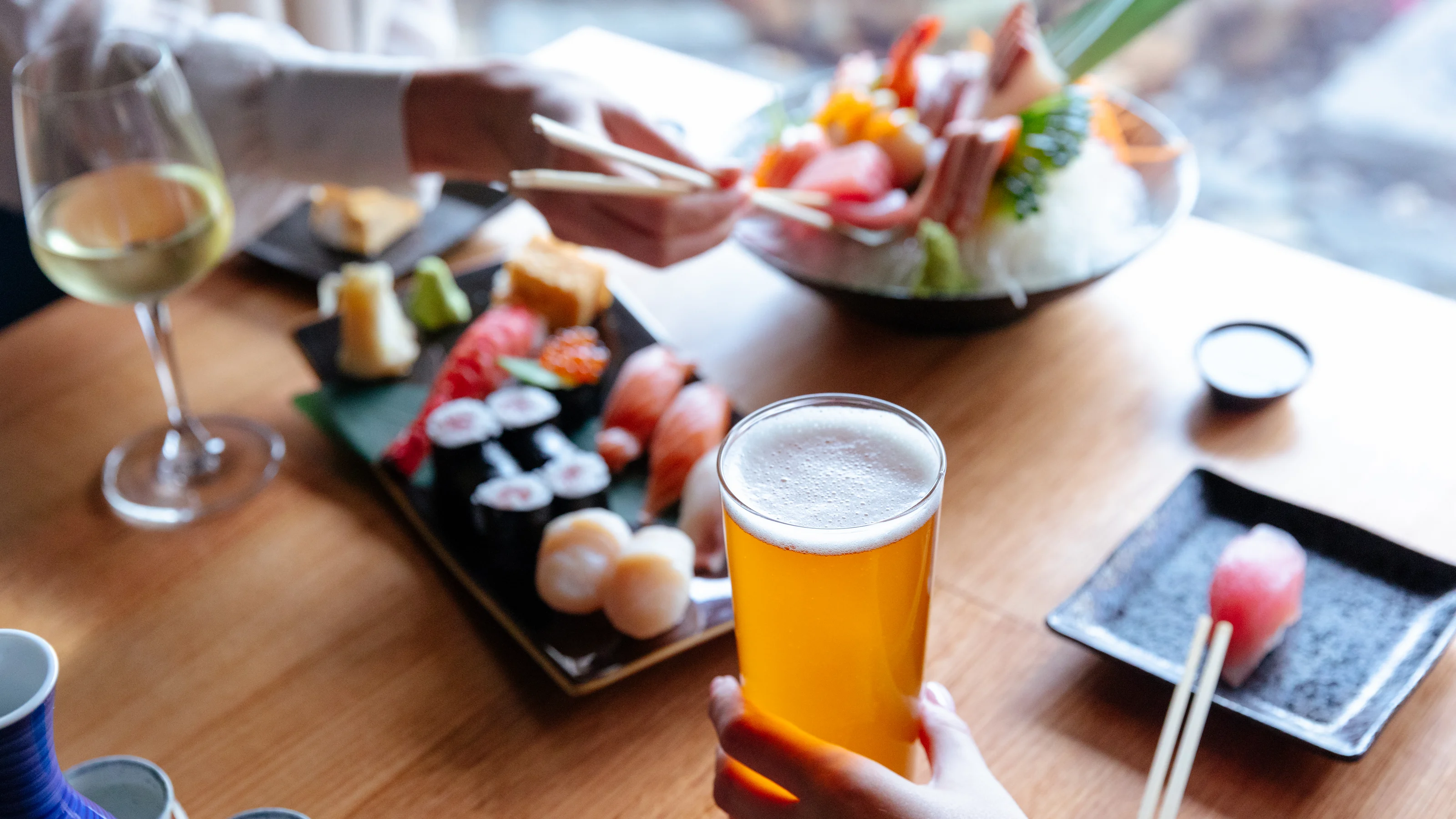 Hands holding chopsticks and a glass of beer over a table at Hatsuhana Japanese Restaurant, with sushi, sashimi, a glass of white wine, and a small dish.