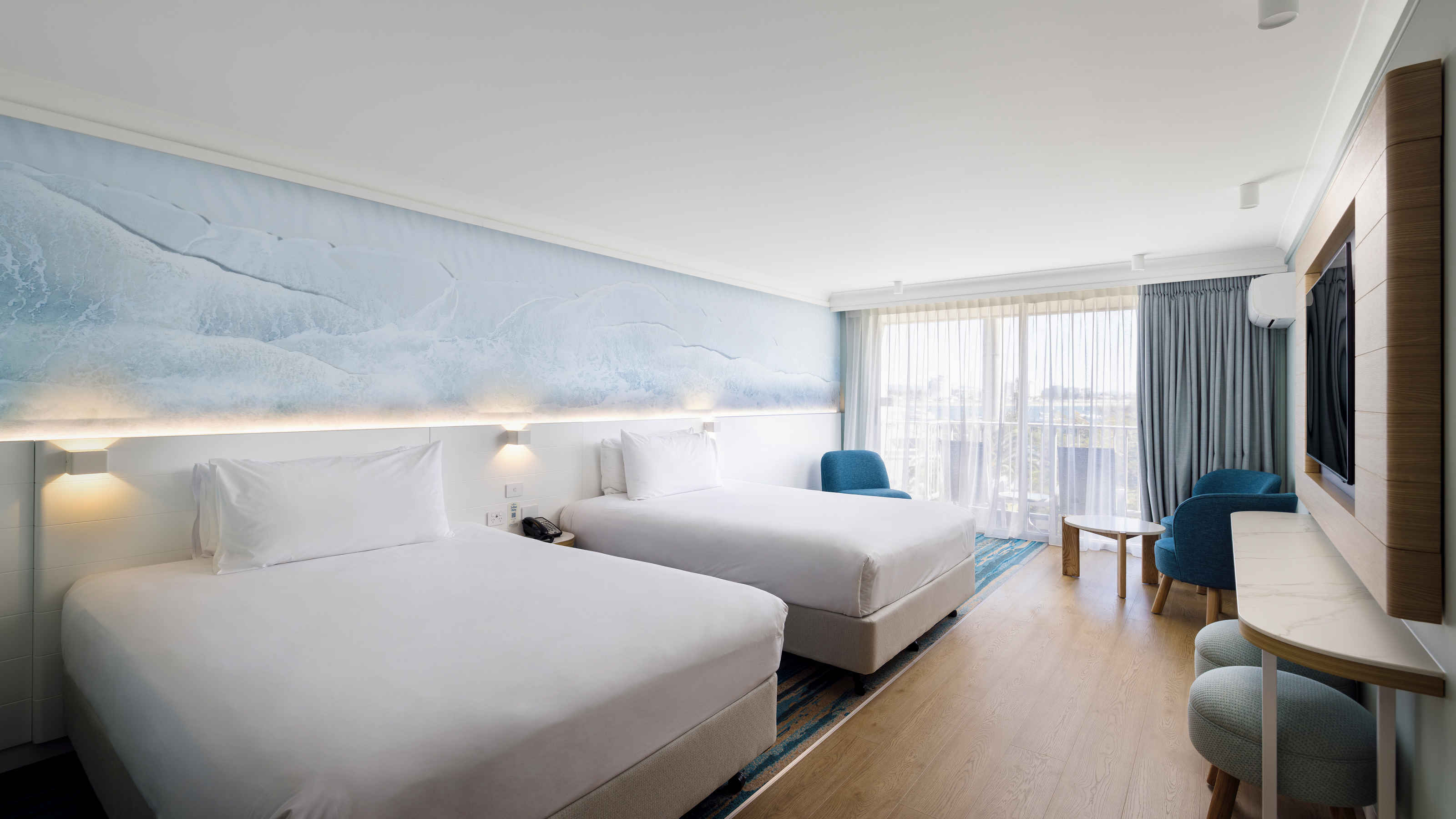 Modern hotel room with two double beds, a large ocean-themed mural, TV, two blue chairs, and a round table. A window with sheer curtains shows a view of a balcony and distant scenery.