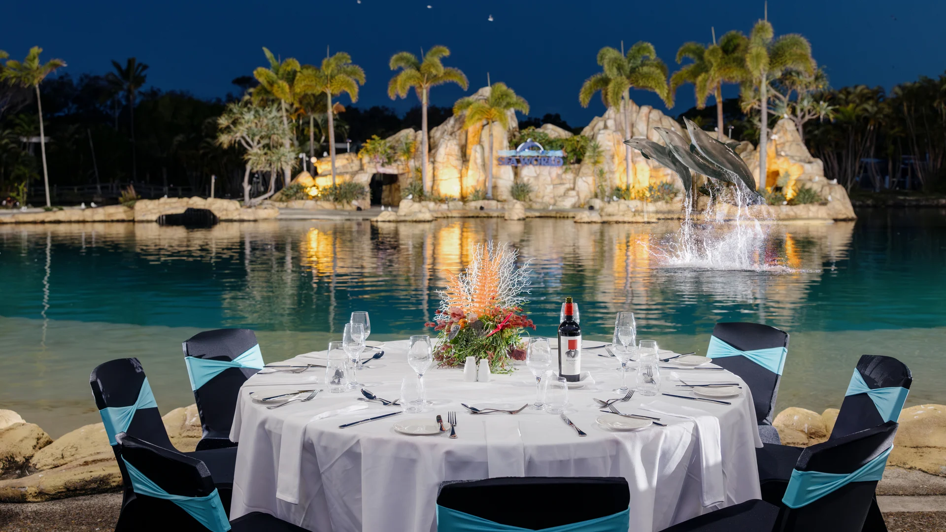 A round table set for dinner with a white tablecloth and wine bottle overlooks a lit water feature with palm trees and dolphins at night.
