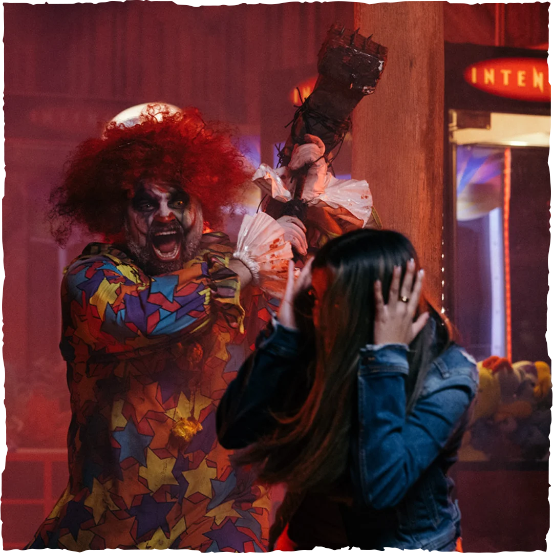 A person dressed as a menacing clown with red hair and colorful costume raises a weapon behind a frightened woman who is covering her face with her hands in fear, inside an arcade with dim, dramatic lighting.