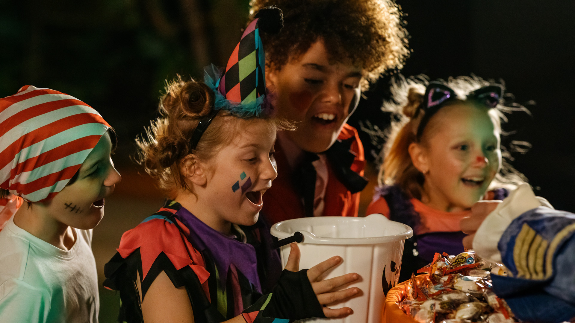 Four children in colorful Halloween costumes excitedly look at a container filled with candy, holding trick-or-treat buckets. They appear happy and are illuminated by warm lighting, likely during nighttime festivities.