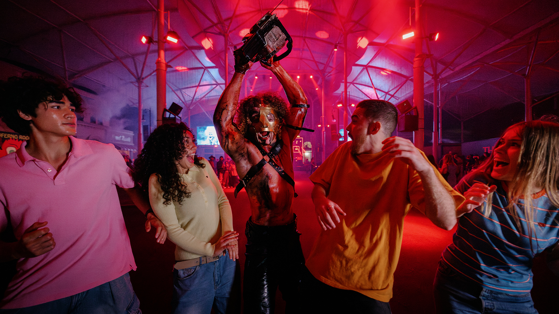 A performer in costume holding a chainsaw poses with four startled people under red lights in a large indoor venue, creating a dramatic and intense atmosphere.